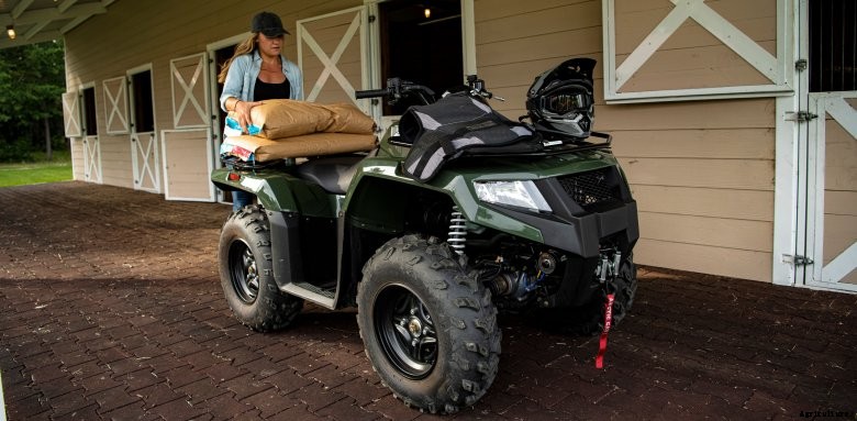 A woman loads horse feed on the back of a green Arctic Cat Altera 450 in front of a tan barn